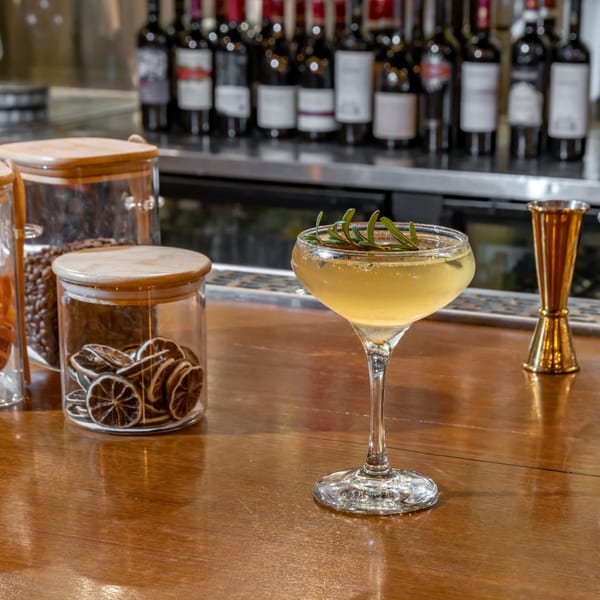 A cocktail garnished with rosemary on a wooden bar, with wine bottles and dried citrus in the background.