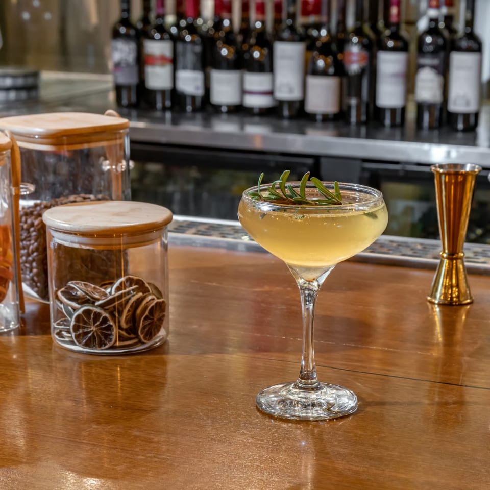 A cocktail garnished with rosemary on a wooden bar, with wine bottles and dried citrus in the background.