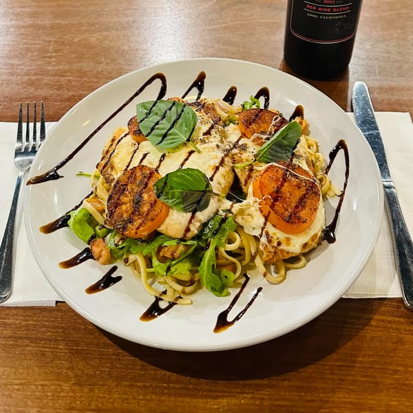 Italian pasta dish with grilled chicken, tomatoes, mozzarella, basil, and balsamic glaze, next to a wine bottle.