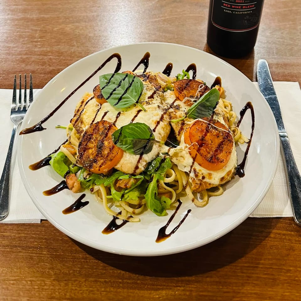 Italian pasta dish with grilled chicken, tomatoes, mozzarella, basil, and balsamic glaze, next to a wine bottle.