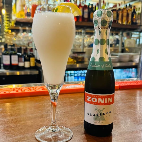 A tall glass of frozen limoncello with a lemon slice next to a Zonin Prosecco bottle.
