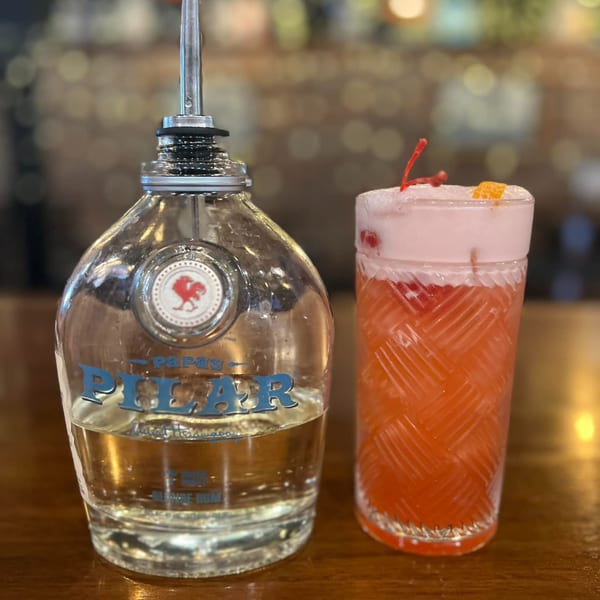 A bottle of Papa's Pilar rum next to a vibrant pink cocktail garnished with a cherry and orange peel.