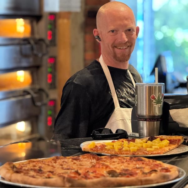 Smiling Bella Napoli Pizzeria chef with two pizzas, one with pineapple, and a hot oven in the background.