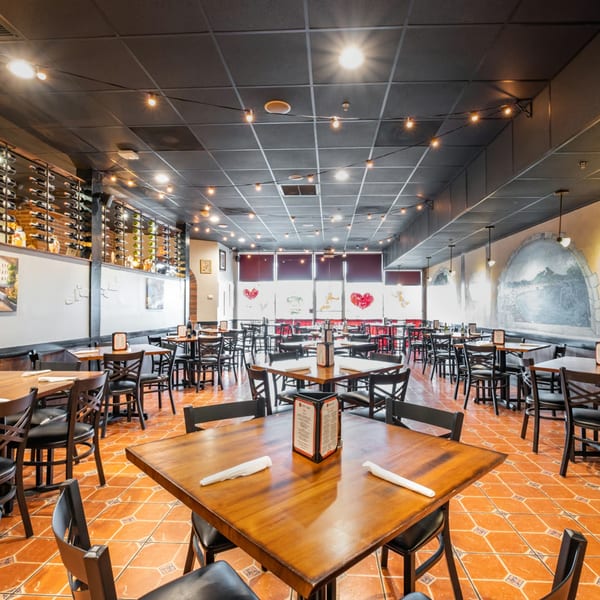 Spacious Bella Napoli Pizzeria dining room with wooden tables, black chairs, wine racks, and string lights.