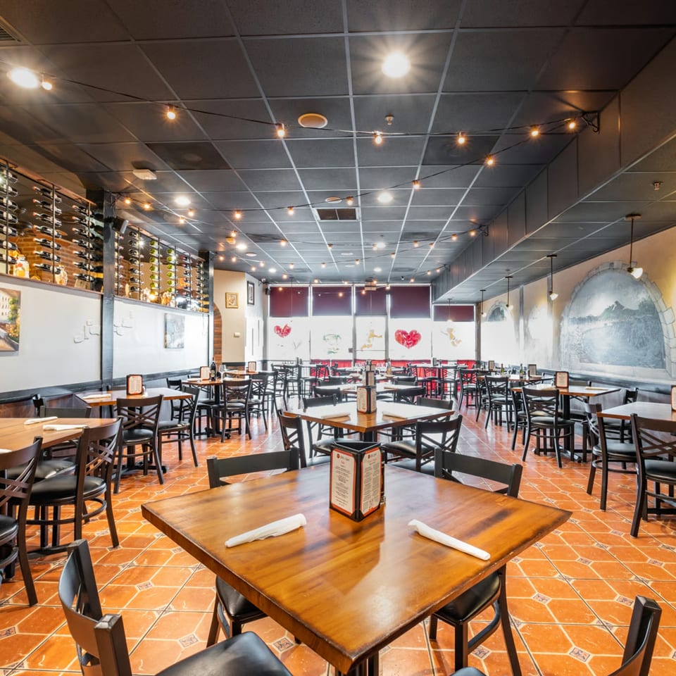 Spacious Bella Napoli Pizzeria dining room with wooden tables, black chairs, wine racks, and string lights.