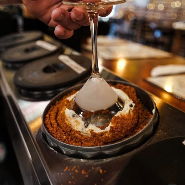 A hand holds a martini glass with dry ice over a dessert in a metal tin.