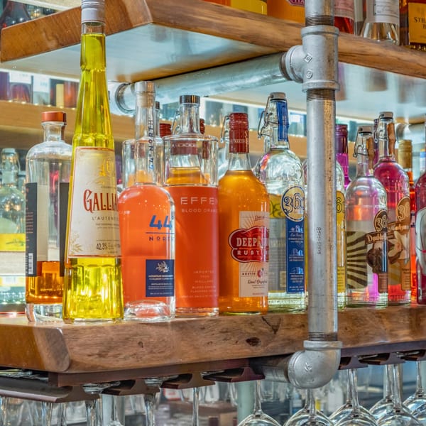 A close-up of various colorful liquor bottles on a wooden shelf with hanging wine glasses.