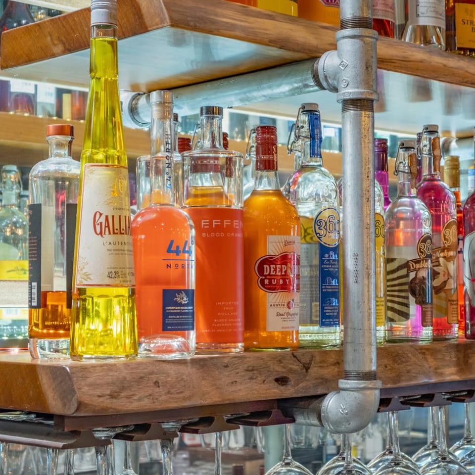 A close-up of various colorful liquor bottles on a wooden shelf with hanging wine glasses.