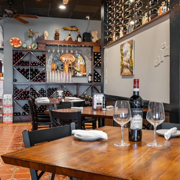 Bella Napoli Pizzeria & Restaurant interior with wine bottles, glasses, and wooden tables set for dining.