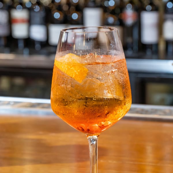 A vibrant orange Aperol Spritz cocktail with ice and an orange slice, on a wooden bar.