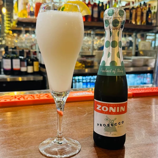 A tall glass of frozen limoncello with a lemon slice next to a Zonin Prosecco bottle on a bar.