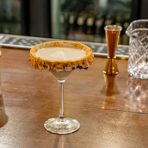 A creamy cocktail in a martini glass with a crushed cookie rim sits on a wooden bar.
