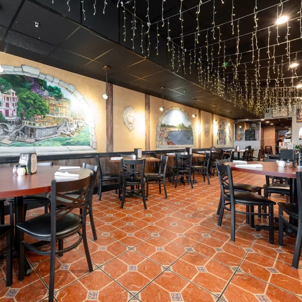 Empty restaurant dining room with murals of Italian coastal towns, string lights, and black tables and chairs.