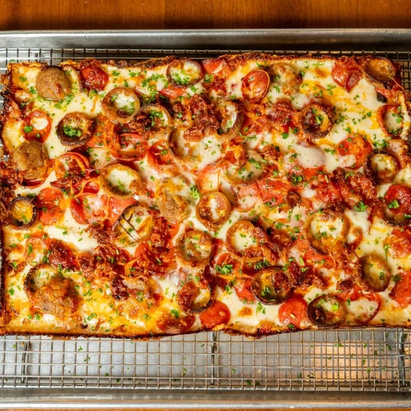 A rectangular Detroit-style pizza with pepperoni, mushrooms, and crispy onions on a metal cooling rack.
