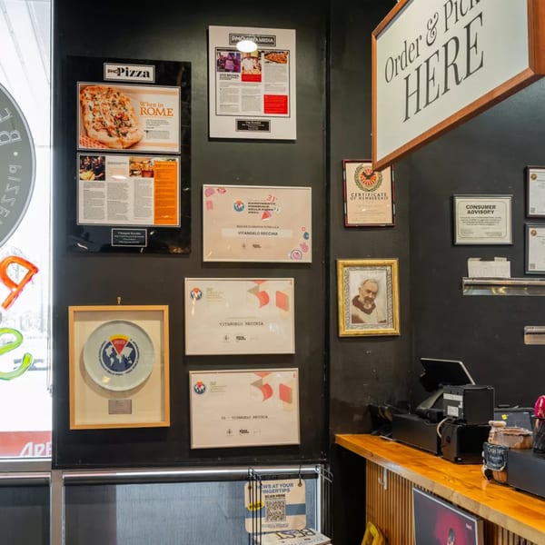 A restaurant wall displaying framed awards, certificates, and a pizza advertisement above a counter.