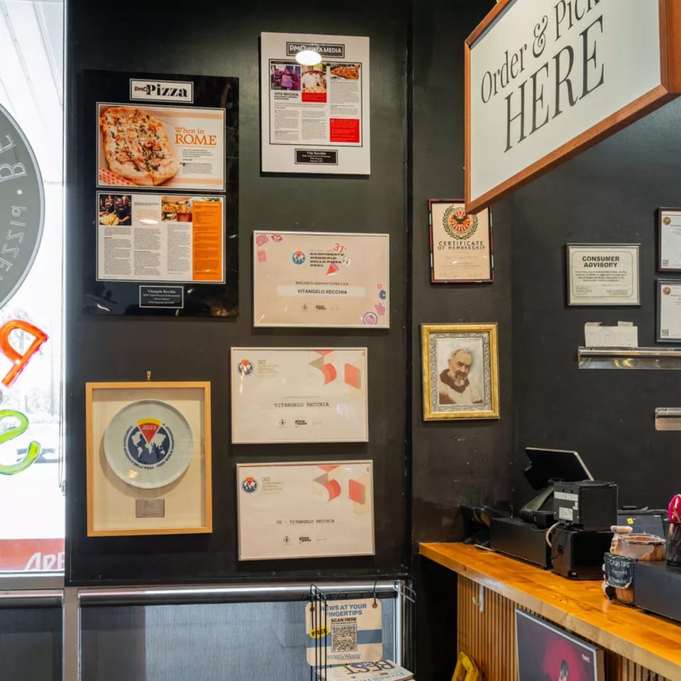 A restaurant wall displaying framed awards, certificates, and a pizza advertisement above a counter.