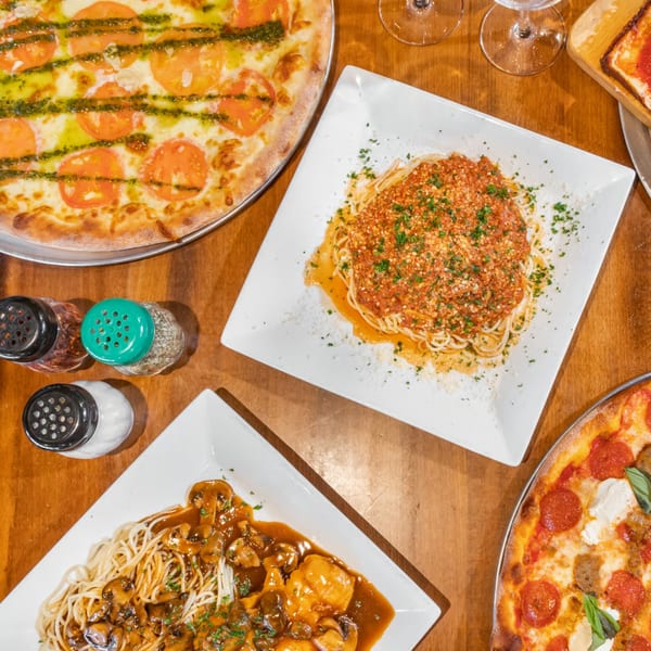 Overhead view of two pizzas and two pasta dishes on a wooden table at Bella Napoli Pizzeria.