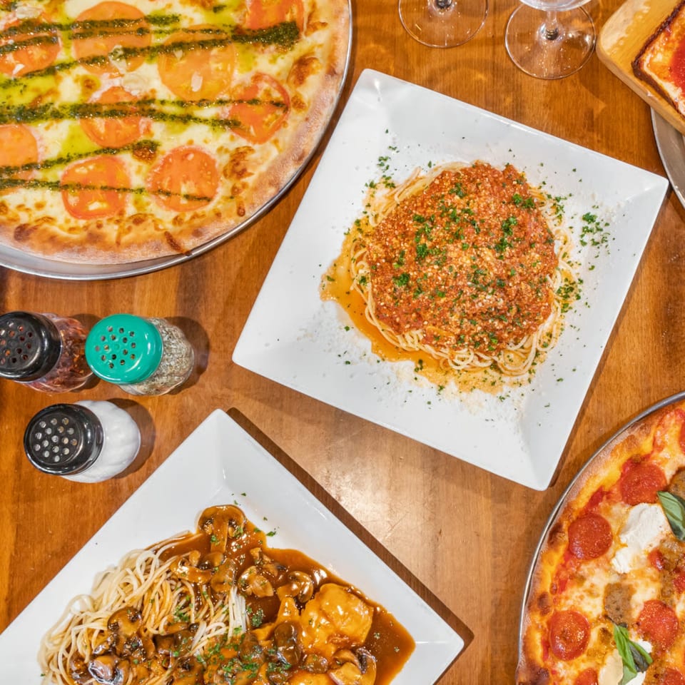 Overhead view of two pizzas and two pasta dishes on a wooden table at Bella Napoli Pizzeria.