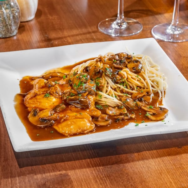 Chicken Marsala with mushrooms and spaghetti on a white plate, with wine glasses in the background.