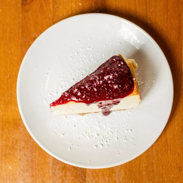 A slice of cheesecake with red berry topping and powdered sugar on a white plate.