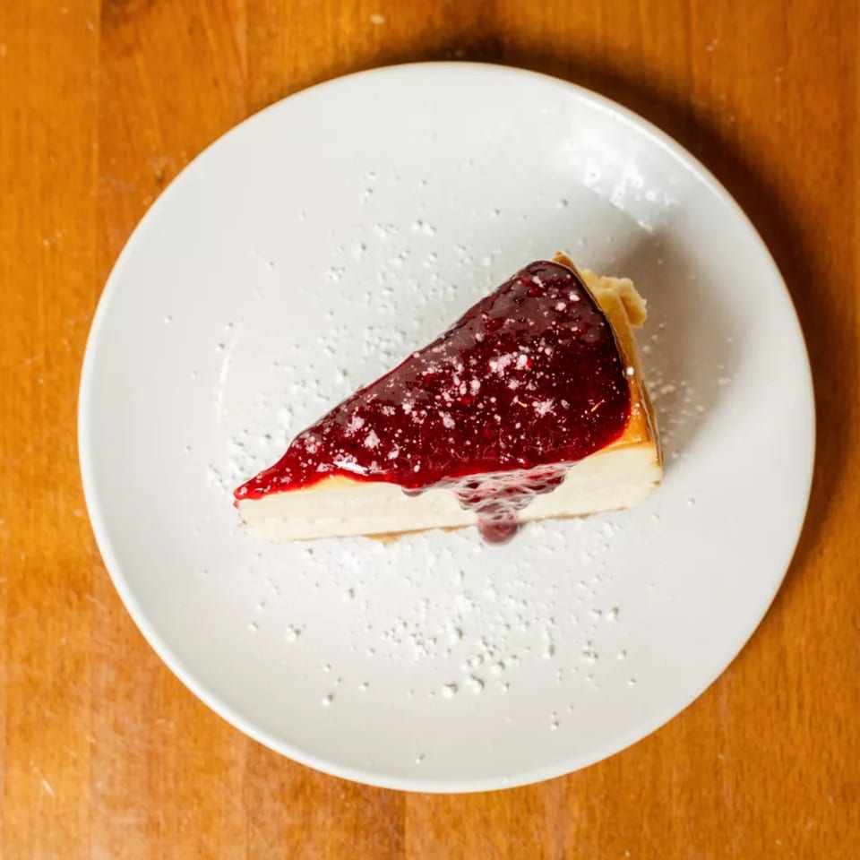 A slice of cheesecake with red berry topping and powdered sugar on a white plate.