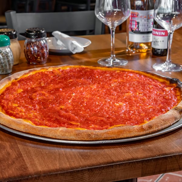 A large, round Bella Napoli pizza with bright red tomato sauce on a wooden table with wine.