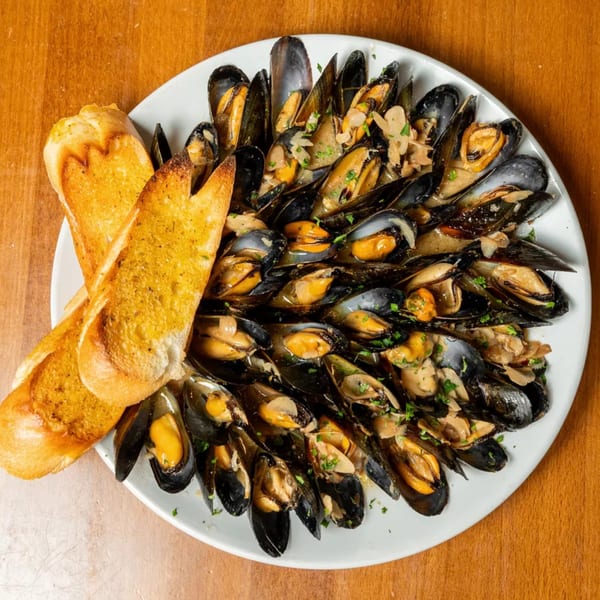 A white plate filled with steamed mussels in a creamy sauce, garnished with parsley and two slices of garlic bread.