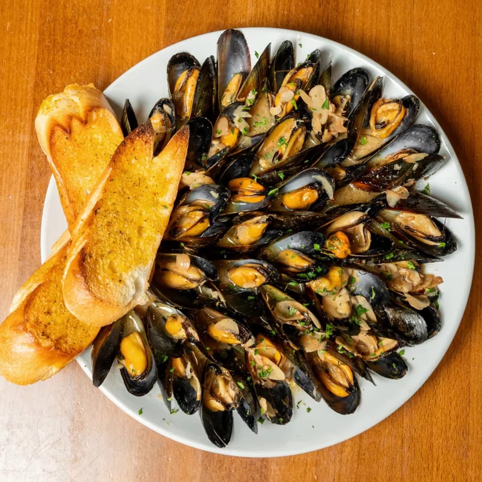 A white plate filled with steamed mussels in a creamy sauce, garnished with parsley and two slices of garlic bread.